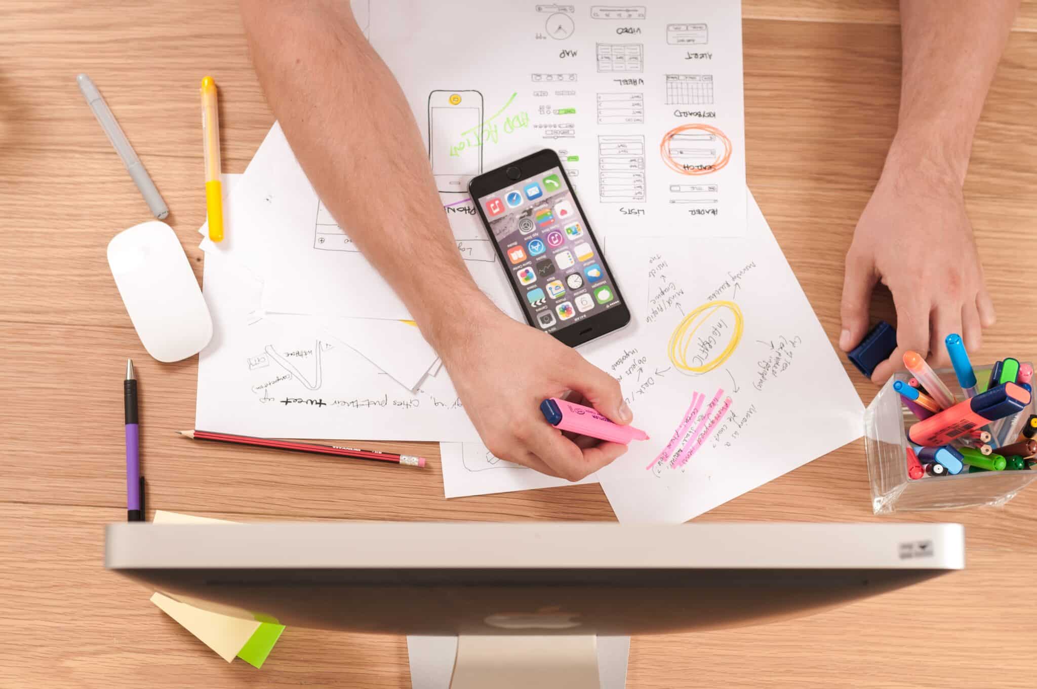 a desk with work papers, and a cellphone in front of a computer screen
