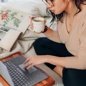 Women drinking coffee, sitting on a bed working on her laptop.