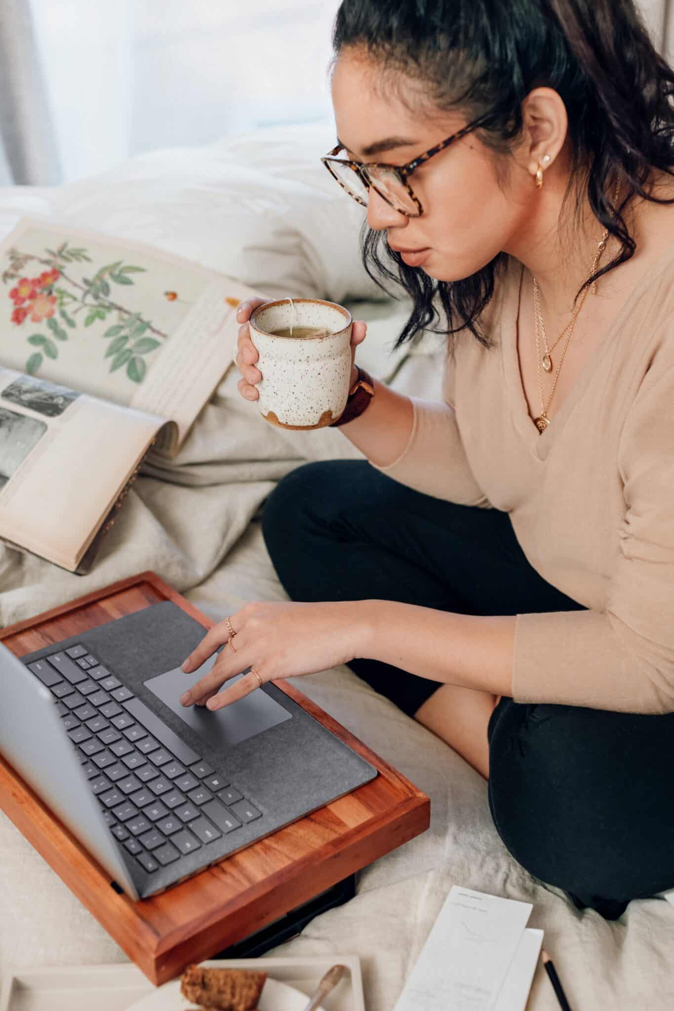 Women drinking coffee, sitting on a bed working on her laptop.