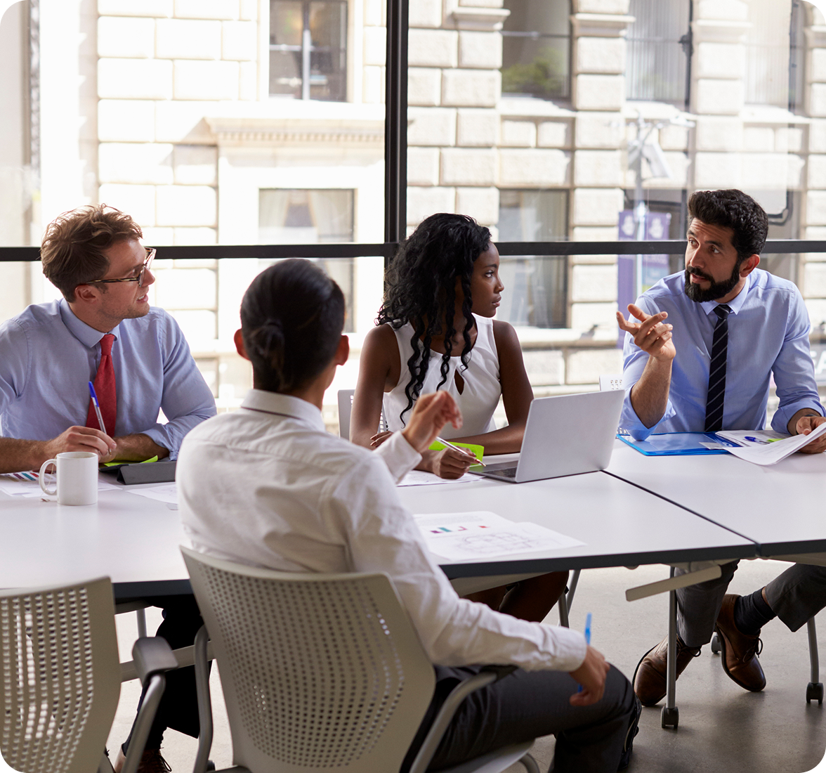 Four people sit around a table in a bright office, engaged in a discussion. One man gestures while speaking; the others listen attentively. Papers, a laptop, and a mug are on the table. Large windows show buildings outside.